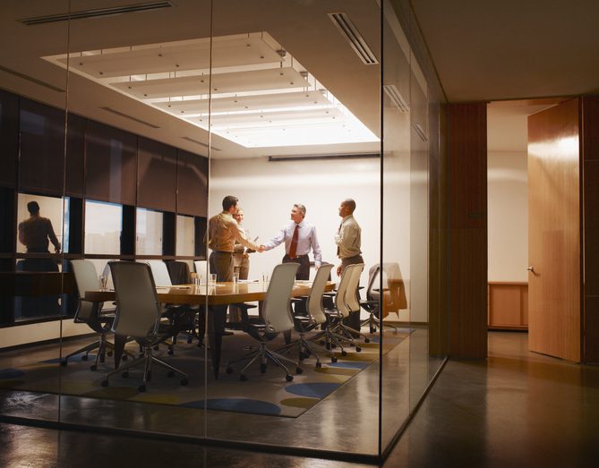 Three people discuss work in a modern glass-walled conference room.